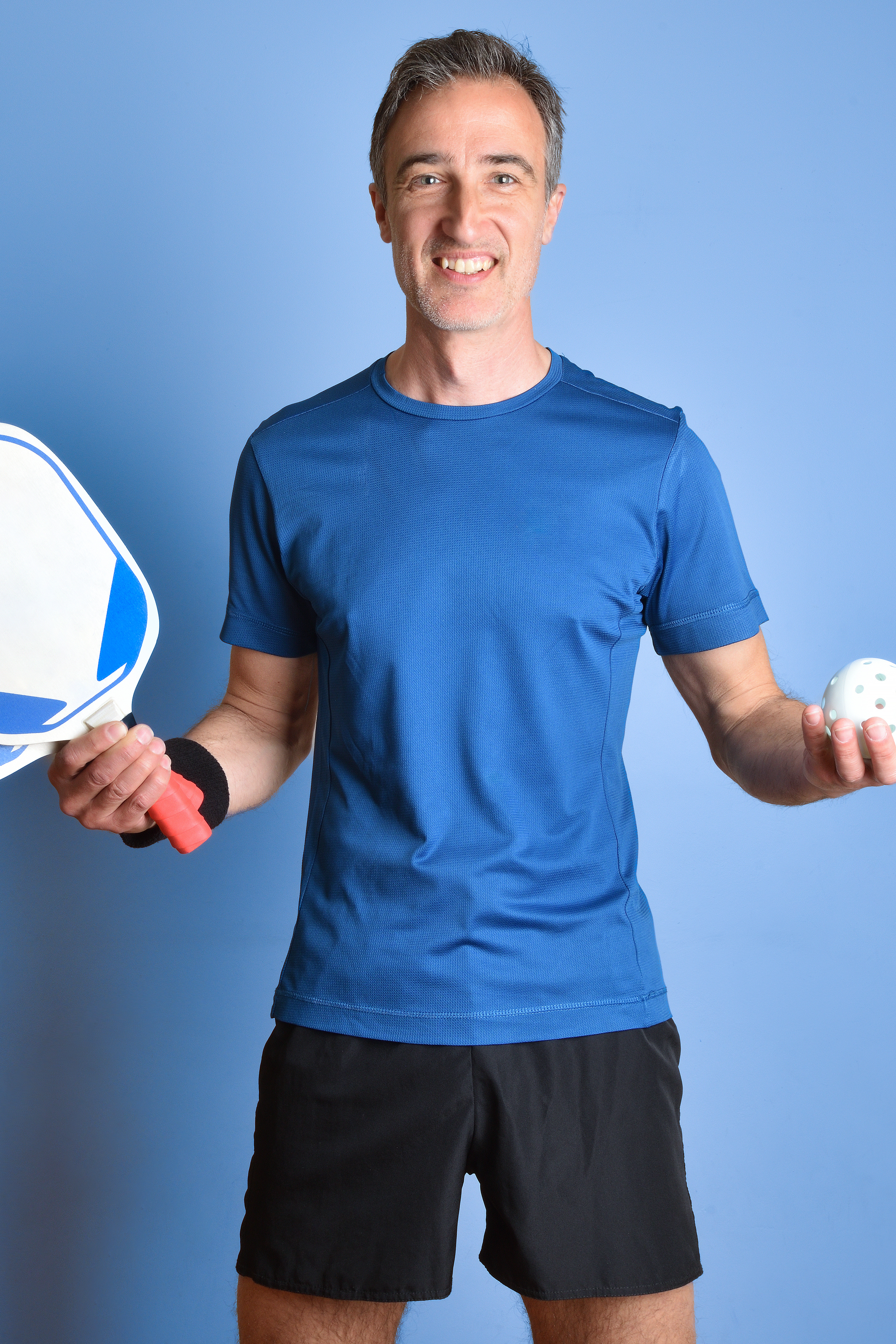 Portrait of pickleball coach Mike MacDonnell smiling in a blue shirt while holding a paddle and ball