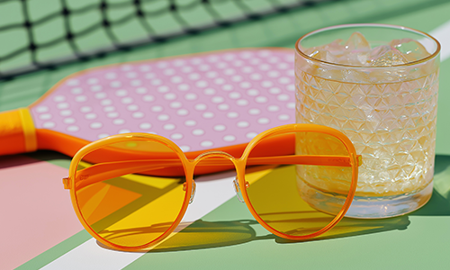 A pink pickleball paddle, yellow sunglasses, and an iced cocktail representing social events