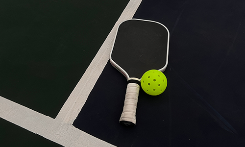 A black pickleball paddle and a yellow pickleball resting on the white boundary line of an indoor court