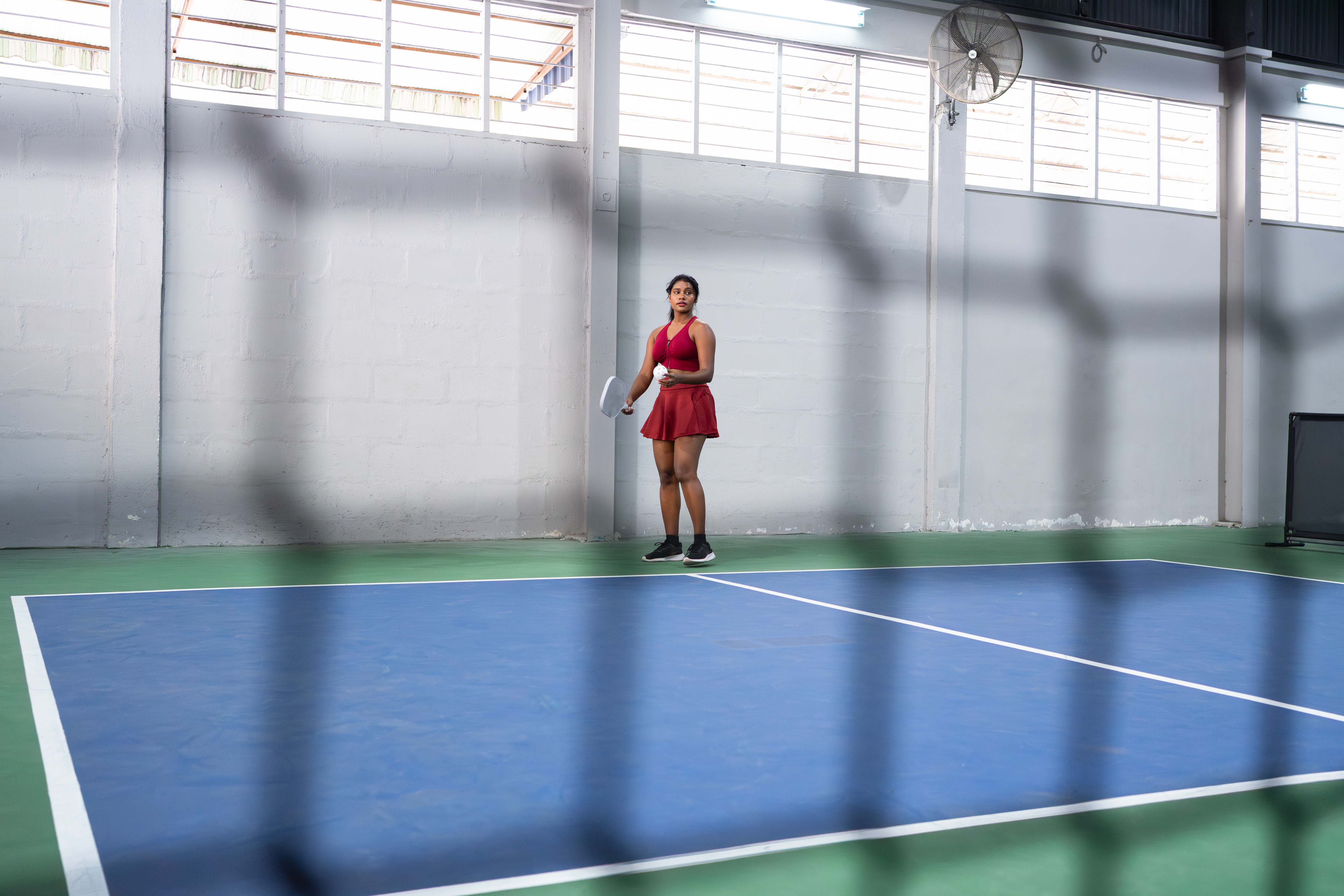 A woman in a red athletic outfit standing on a blue indoor pickleball court viewed through the net