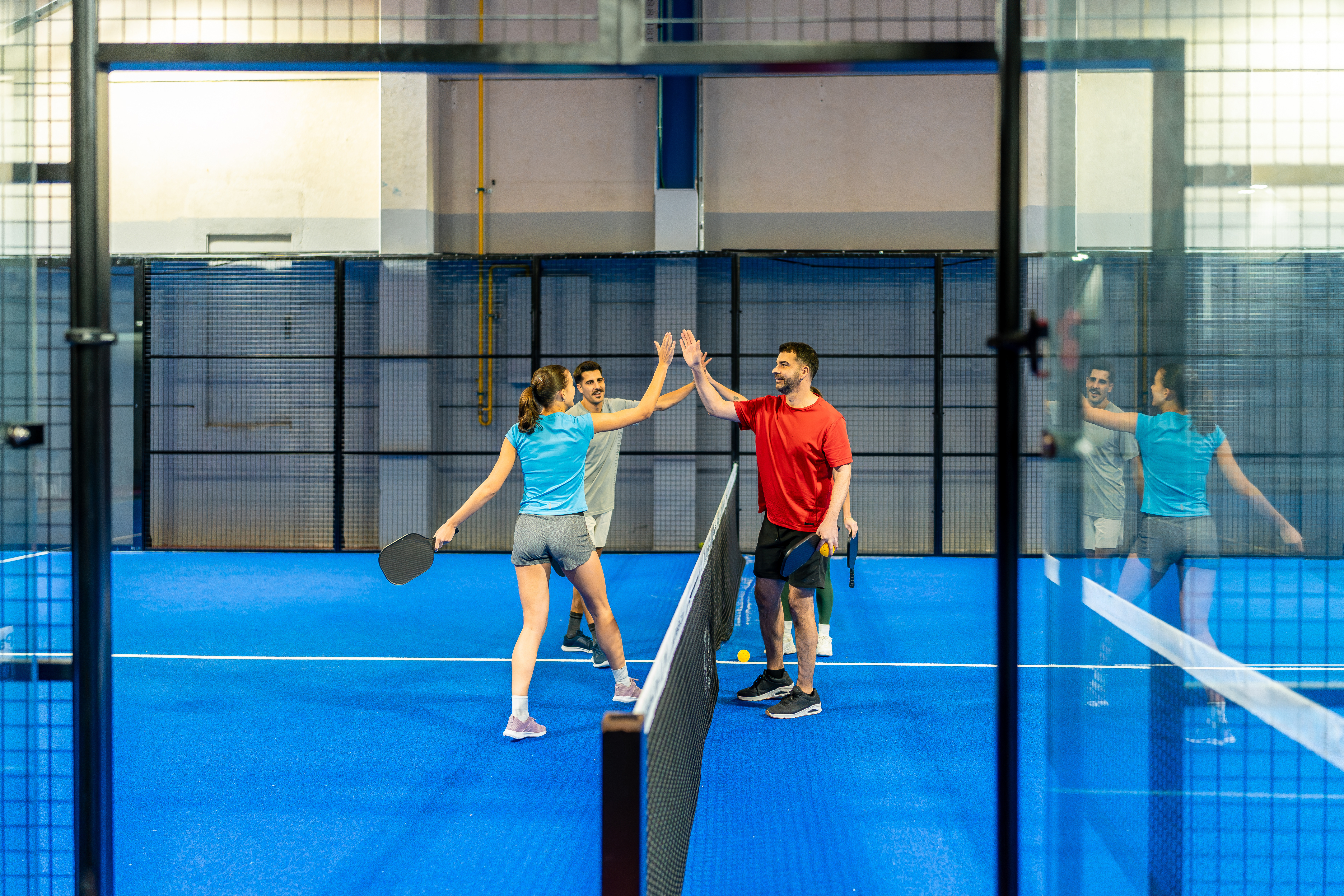 Four pickleball players high-fiving at the net after a doubles match on a blue indoor court