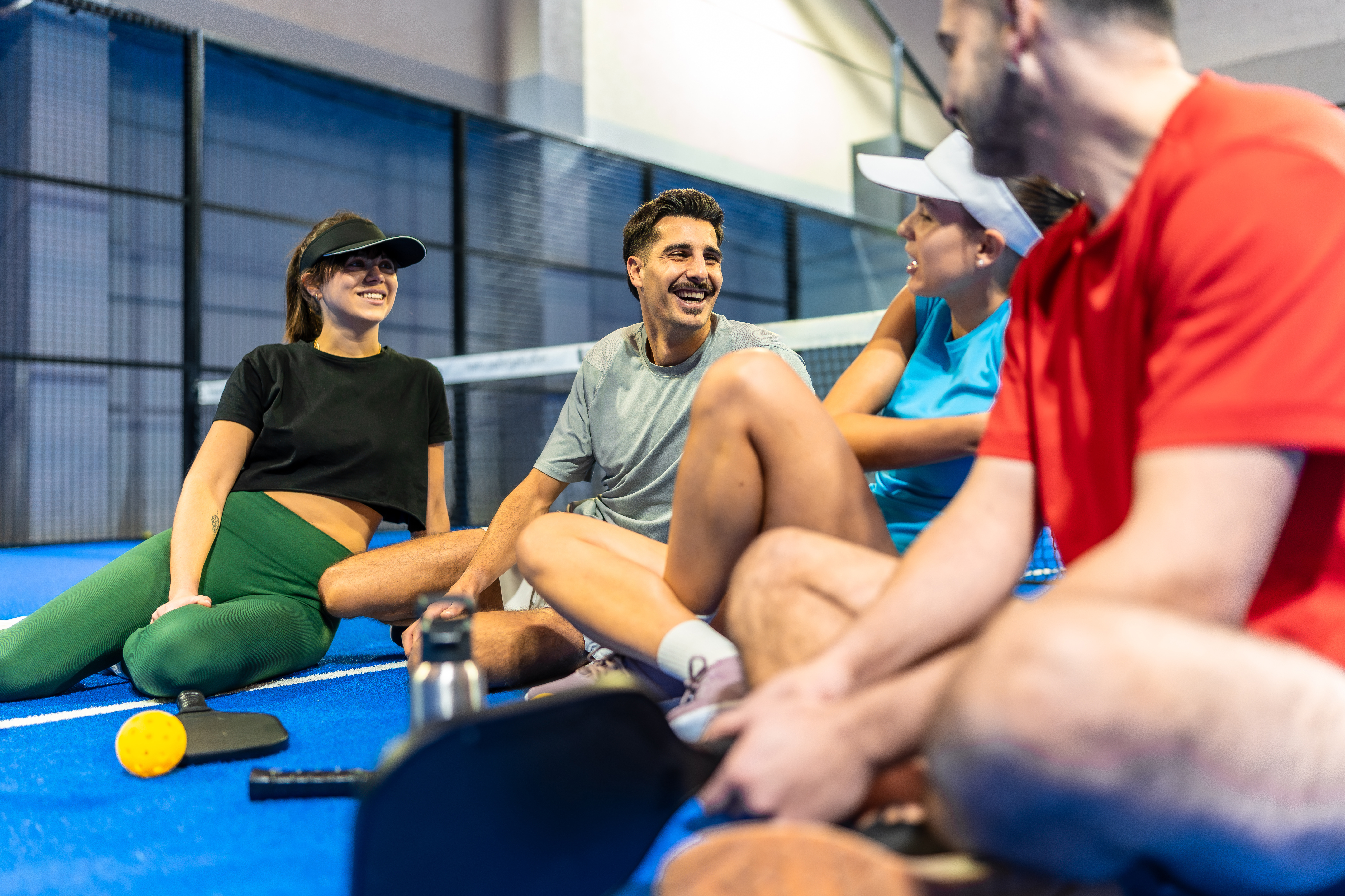 Four pickleball players sitting on a blue indoor court and laughing together after a match