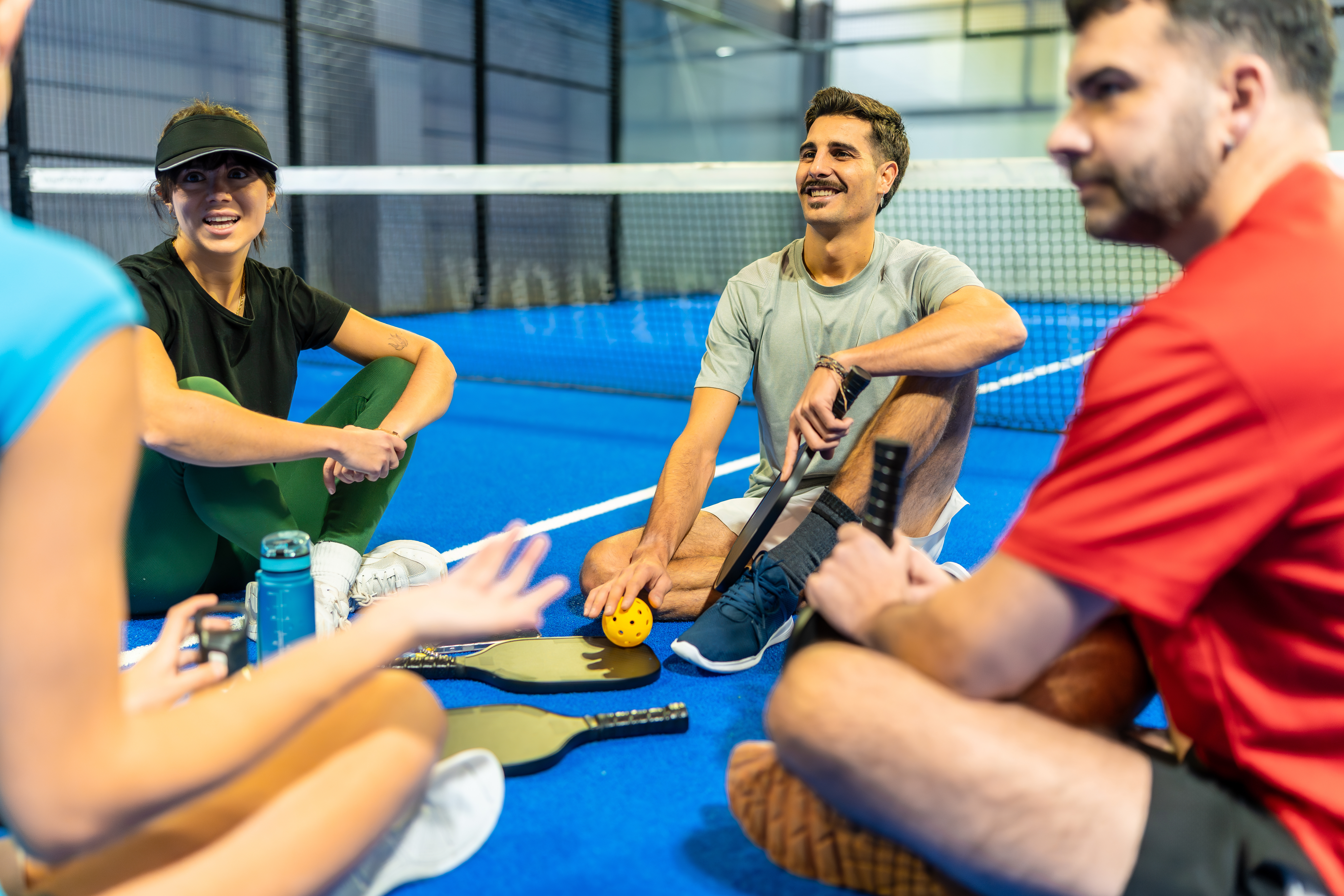 A group of pickleball players sitting on the court talking, with a smiling man holding a paddle and yellow ball